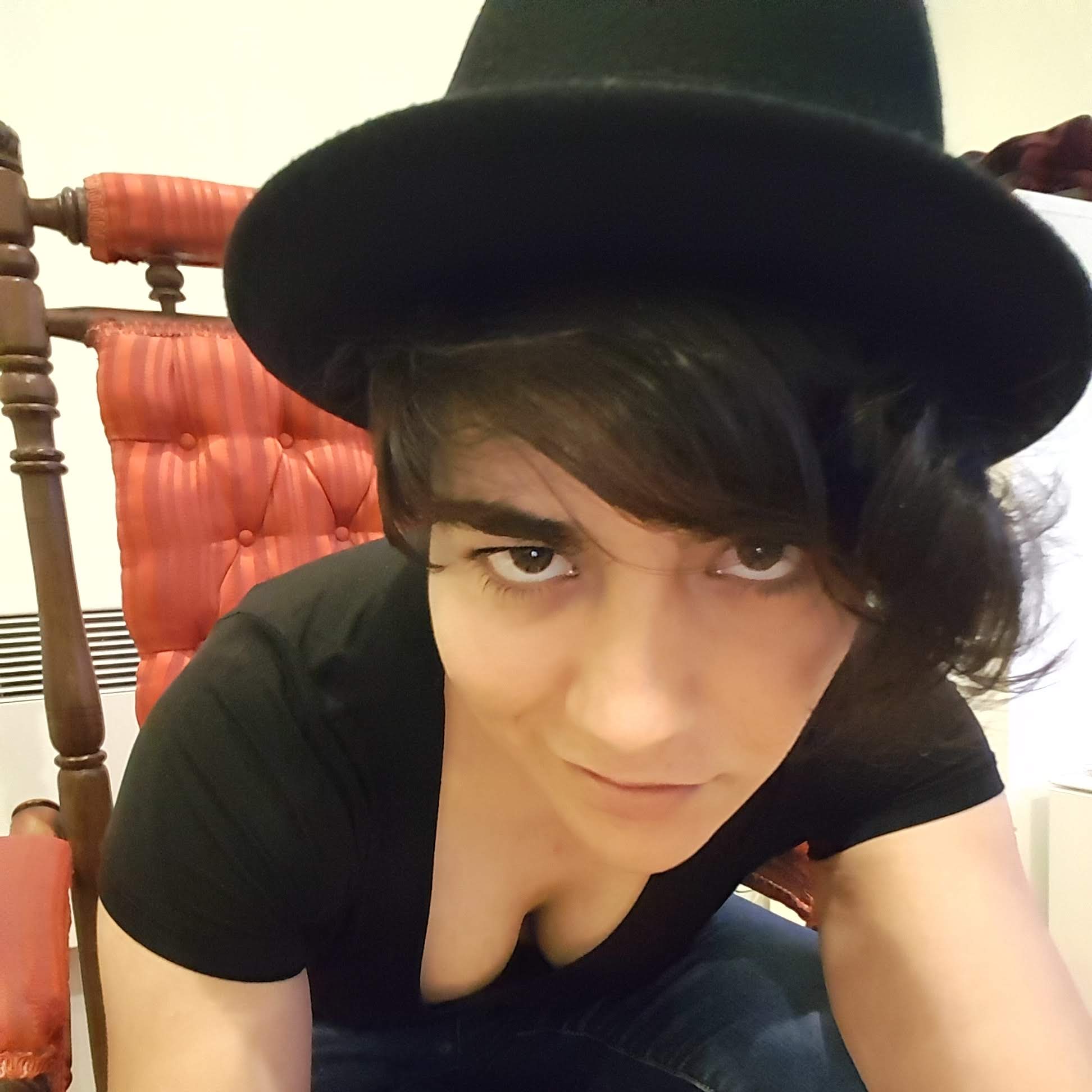 A sly-looking person in a black bowler hat and low-cut top leans forward in their red throne-like chair with a smirk on their face.